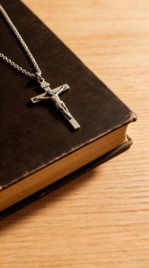 Yakın plan, yüksek açılı gümüş bir haç kolyesinin, eski, koyu renkli kutsal bir İncil 'in üzerine örtülmüş, sıcak ahşap bir arka planda çekilmiş fotoğrafı. Sahne, inanç, maneviyat ve dini bağlılığı çağrıştırıyor..