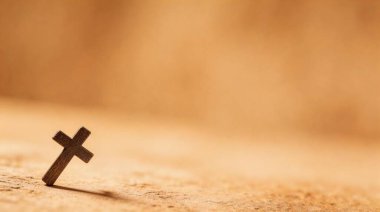 Küçük, basit bir ahşap haç desenli kahverengi bir yüzey üzerinde dimdik durur, ince bir gölge bırakır. Arkaplan bulanık, metin veya diğer tasarım elemanları için yeterli kopya alanı sağlıyor. Bu görüntü inanç, maneviyat ve umudu çağrıştırıyor..
