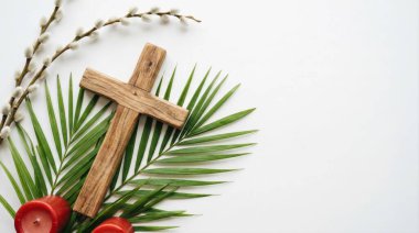 A flat lay composition featuring a wooden Christian cross, green palm fronds, pussy willow, and red candles on a white background with copy space for text.