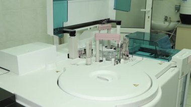 Steril bir klinik laboratuvarında modern bir robot analizcinin detaylı görüntüsü. Sistem, yüksek hassasiyetli biyokimyasal araştırma ve tanı testleri için otomatik silahlar ve örnek atlıkarıncalardan oluşuyor..