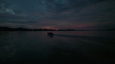 Alacakaranlıkta sakin denizdeki küçük motorlu tekne, pembe ufuk ve karanlık dağ siluetleri ile dramatik bulutlu gökyüzü altında uyanır; Koh Yao Yai, Tayland 'da sakin karamsar deniz manzarası.