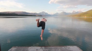 Arkasında panoramik dağlar olan el değmemiş bir göle dalan genç adam. McDonald Gölü Buzul Ulusal Parkı, Montana.
