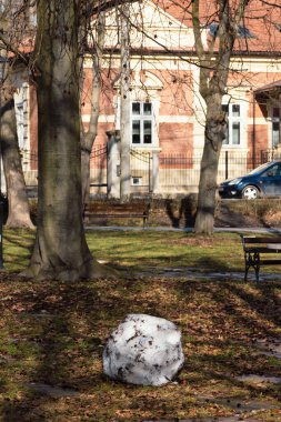 Büyük, kirli bir kartopu çimenli bir parkta kış sona ererken ve bahar başlarken duruyor. Arka planda çıplak ağaçlar, banklar ve kentsel bir bina mevsimsel geçiş, erime ve iklim değişikliği kavramını vurguluyor.