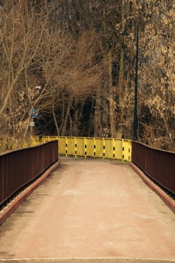 Boş yaya köprüsü geçidi, kırmızı parmaklıklar, sarı güvenlik bariyerleri, bir sokak lambası, ve sakin bir kış gününde arka planda yapraksız ağaçlar..