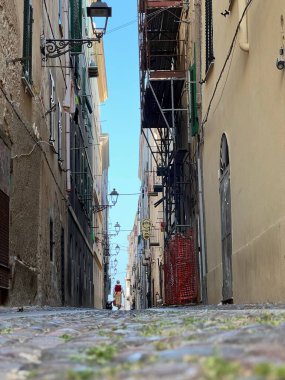 Taş kaldırımları, tarihi binaları ve sokak lambaları olan dar bir kasaba sokağının alçak açılı görüntüsü. Uzakta yürüyen bir kadın otantik bir Akdeniz şehir atmosferi yaratır..