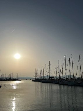 Sakin bir yat limanında gün batımı sisli. Rıhtım boyunca yelkenliler dizilmiş. Güneş ışığı, pürüzsüz suya yansıyarak, huzurlu bir minimalist kıyı seyahati sahnesi oluşturur..