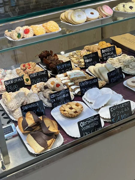 İtalyan pastanesi vitrininde çeşitli geleneksel kurabiyeler, tepsilerde pastalar, el yazısı fiyat tabelaları, bir pastanenin vitrininde taze tatlılar..