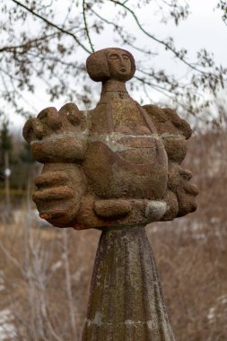 Soyut insan yüzüne sahip uzun, yıpranmış bir halk sanatı heykeli ve el benzeri şekiller, açık havada yumuşak bulanık kış arka planına ve bulutlu gökyüzüne karşı fotoğraflanmış..