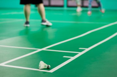 Badminton kortunda bulanık oyuncularla yeşil katta beyaz badminton topu. Spor konsepti. Badminton topu (servis horozu), badminton sahası oyuncuları ile arka planda modern spor salonu, seçici odak noktası. 