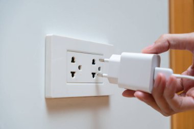 Bir kadına beyaz bir güç adaptörünü duvar prizine bağlayan bir el yaklaş. Şarj cihazları ve elektrik tüketimi kavramı.