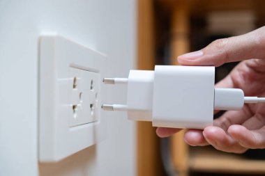 Asyalı bir kadın beyaz bir adaptörü duvar prizine takıyor. Cihazları şarj etme ve evde elektrik kullanma kavramı.
