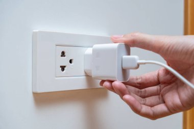 Bir kadına beyaz bir güç adaptörünü duvar prizine bağlayan bir el yaklaş. Şarj cihazları ve elektrik tüketimi kavramı.