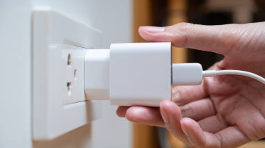 Bir kadına beyaz bir güç adaptörünü duvar prizine bağlayan bir el yaklaş. Şarj cihazları ve elektrik tüketimi kavramı.