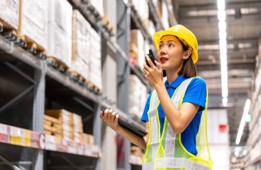 Güvenlik kaskı ve yansıtıcı yelek giyen güzel Asyalı mühendis kadın raflardaki malzeme ve malzemeleri tabletle kontrol ediyor ve depoda telsiz iletişimi olan biriyle konuşuyor..