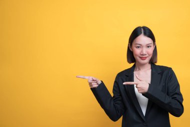 Sarı arka planda siyah takım elbiseli güzel bir Asyalı kadın sol tarafı işaret ediyor ve gülümsüyor..