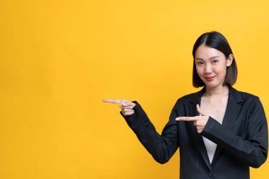 Sarı arka planda siyah takım elbiseli güzel bir Asyalı kadın sol tarafı işaret ediyor ve gülümsüyor..