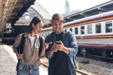 Mutlu Asyalı yakışıklı adam ve güzel kadın sırt çantalı sırt çantalı sırt çantalı bir adam tren istasyonunda birlikte yürüyor ve tren saatlerini cep telefonuyla kontrol ediyor..