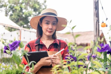 Portre mutlu Asyalı kadın bahçıvan şapkalı ve kahverengi önlüklü çiçek tarhını tabletle kontrol ediyor ve gülümsüyor çünkü yeşil evde ekimi başarılıydı..