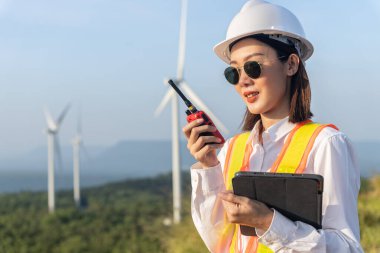 Güvenlik kaskı ve yansıtıcı yelek giyen güzel Asyalı kadın mühendisin portresi katlanır kollar ve rüzgar türbininin arka planında telsiz tutuyor. Çevre dostu.
