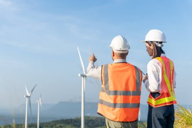Güvenlik kaskı takan Asyalı erkek ve kadın mühendis ve yansıtıcı yelek takımının rüzgar türbini pervane arka planıyla çalışmalarının arka görüntüsü. Temiz enerjinin geleceği için çevre dostu.