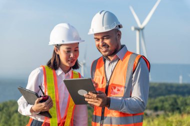 Gülümseyen güzel Asyalı kadın ve güvenlik kaskı ve yansıtıcı yelek giyen yakışıklı erkek mühendisler arka planda rüzgar türbini pervanesi ve açık mavi gökyüzü olan tabletle çalışıyorlar..