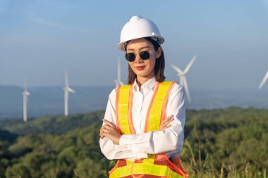 Güneş gözlüğü takan güzel Asyalı kadın mühendisin portresi, güvenlik kaskı ve rüzgar türbini pervaneli yansıtıcı yelek. Alternatif enerji, gelecek için çevre dostu.