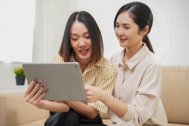 Sevgi LGBT konsepti. Mutlu Asyalı güzel lezbiyen çift yavaş çekimde oturma odasında konuşuyor ve tablet kullanıyor. Kaliteli zaman, eğlence, teknoloji ve aşk harcamak.