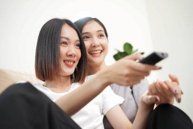 Sevgi LGBT konsepti. Mutlu Asyalı güzel lezbiyen çift, evde kanepede oturup, yavaş çekimde akşam TV izlerken birbirlerine sarılıp gülüyorlar. Kaliteli zaman geçirme.