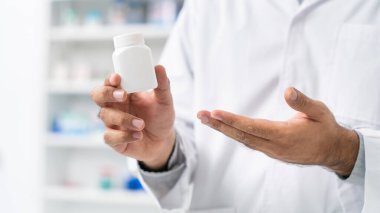 Beyaz laboratuvar önlüklü Asyalı bir eczacının, elinde bir ilaç şişesiyle modern eczanenin raflarındaki bulanık eczane raflarına karşı durduğu yakın plan..