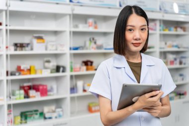 Laboratuvar önlüğü giymiş, elinde tablet olan mutlu Asyalı kadın eczacı kameraya bakıyor ve eczanedeki işinden gurur duyuyor..