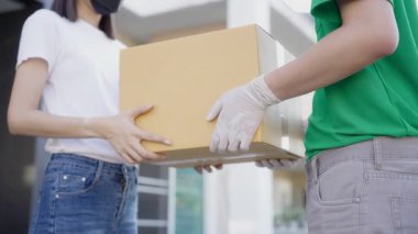 Medium shot. Asian delivery man wearing green uniform, protection mask and medical rubber gloves send a cardboard box to customer and she sign on the phone in front of the customer's house.