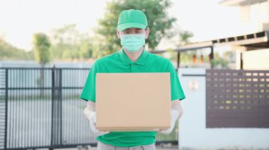 Corona Virüsü konsepti, yeşil üniformalı yakışıklı teslimatçı koruma maskesi ve tıbbi kauçuk eldivenli karton kutu tutuyor. 4k çözünürlüğü.