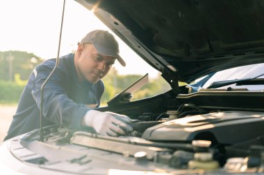 Üniforma ve koruma eldiveni giyen Asyalı otomobil tamircisi. Tabletli, araba servisli ve bakımlı bir araba motoru otomotiv atölyesini tamir ediyor..
