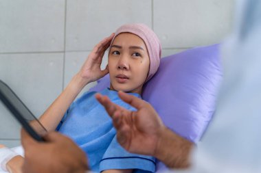 Asyalı kadın hasta semptomlarını bir onkolojiste yatakta otururken ve hastane koğuşunda laboratuvar önlüğü giymiş bir adama bakarken anlatır..