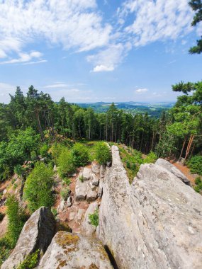 Yeşil yemyeşil ağaçları, kayalık oluşumları ve dağınık taşları olan Çek ormanlarına bakan bir dağın tepesinden dinamik bir manzara. Yumuşak bulutlu parlak mavi gökyüzü canlı doğal manzarayı güçlendirir. Doğa kavramı, macera, yürüyüş.