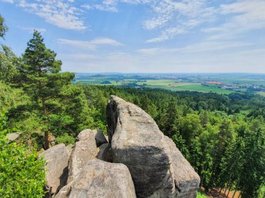 Yeşil yemyeşil ağaçları, kayalık oluşumları ve dağınık taşları olan Çek ormanlarına bakan bir dağın tepesinden dinamik bir manzara. Yumuşak bulutlu parlak mavi gökyüzü canlı doğal manzarayı güçlendirir. Doğa kavramı, macera, yürüyüş.