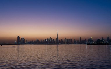 Alacakaranlıkta büyüleyici Dubai silueti: Burç Halife 'nin silueti, sakin sulara yansıyan şehir ışıkları, değişken bir günbatımı gökyüzüne karşı..