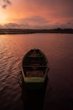 Durgun suda dinlenen yeşil ahşap bir teknenin yer aldığı sakin günbatımı sahnesi, gökyüzünde parıldayan turuncu ve altın tonlarla yansıtılıyor..