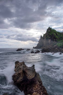 Dalgalar kayalık bir plajda uzun süre rahatça süzülüyor. Bulutlu öğleden sonra ışığı bu sakin kıyı manzarasına dengesiz bir atmosfer ekler..