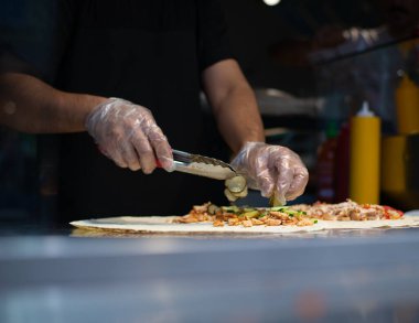 Fast food dükkanında yemek yaparken çekilmiş karanlık atmosferik bir fotoğraf. Tavuk eti pide ekmeğinde kaynatılır. Aşçının eldivenli elleri lezzetli sokak yemekleri yaratıyor. Yüzü yok. Masa ürünleri ve şef.