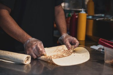 Fast food dükkanında yemek yaparken çekilmiş karanlık atmosferik bir fotoğraf. Tavuk eti pide ekmeğinde kaynatılır. Aşçının eldivenli elleri lezzetli sokak yemekleri yaratıyor. Yüzü yok. Masa ürünleri ve şef.