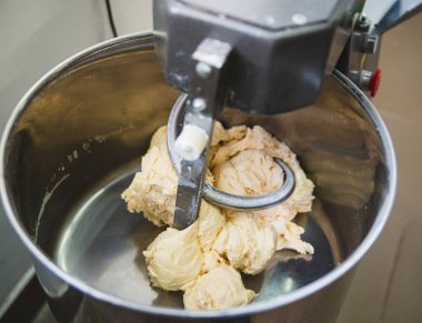 Büyük bir metal kasede yumuşak, hafif bir hamur yoğuran endüstriyel bir mikserin yakın çekimi. Hamuru yoğurma işlemi, mikser kancası merkezde, hamur dizisini aktif olarak karıştırma.