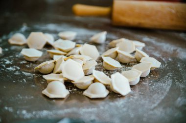 Yakın planda, un ile bir masanın yüzeyine serpilmiş taze mantı tasvir ediliyor. Arka planda ahşap bir merdane var. Fotoğraf aşçılık sanatı ve doğallık atmosferini aktarıyor.