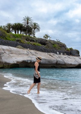 Tropik plajda Rocky Cliff ve Palm Peyzaj 'la çıplak ayakla yürüyen şık bir kadın. Tropik bir plajın kıyılarında yalın ayak yürüyen zarif bir kadın. Yumuşak dalgalar, doğal kaya oluşumları ve palmiye ağaçları sakin bir yolculuk atmosferi yaratır. Mükemmel.