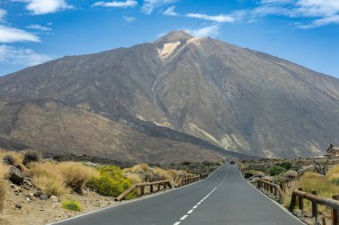 Mavi Gök Altında Açık Asfalt Yolu ile Manzaralı Volkan Manzarası. Parlak mavi gökyüzünün altında düz bir asfalt yolda yükselen dramatik volkanik dağ. Doğal dokular ve geniş perspektif güçlü bir seyahat ve özgürlük kavramı yaratır. Mükemmel.