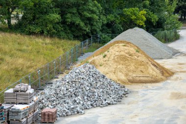 Organize kum, çakıl ve kaldırım taşı yığınları olan endüstriyel Consdoor inşaat deposu. Paletler kaldırım taşları yığılmış ve peyzaj veya altyapı projeleri için hazır. Doğal yeşil geçmişi olan endüstriyel malzeme sahnesi