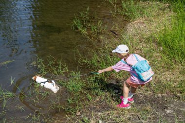Küçük kız, Sunny Nature Setting 'deki nehir kıyısında küçük bir köpekle oynuyor. Küçük köpeği suda yüzerken bir nehrin kenarında duran genç kız. Parlak yaz ışığı ve yeşil çimenler dışarıda huzurlu bir atmosfer yaratır. Çocukluk kavramı