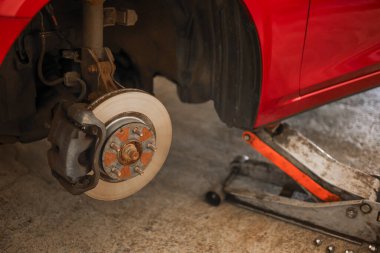 Bir oto tamirhanesinde fren yaparken tekerleği sökülmüş bir araba. Görünür fren diski ve kalibre mekanik onarım ve araç bakım işlemini vurguluyor. Otomotiv endüstrisi, güvenlik denetimi, ulaşım hizmetleri ve
