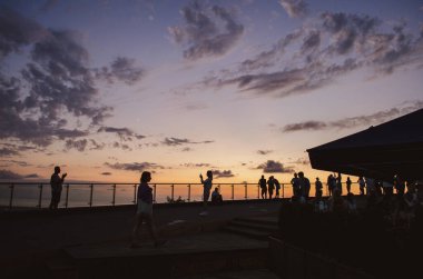 Gökyüzünde hilal şeklinde bir ay olan güzel bir gün batımını terastan izleyen insanların siluetleri..