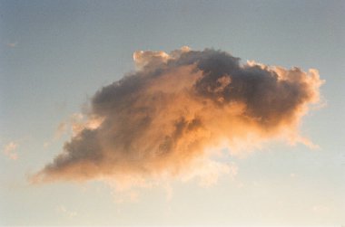 Sıcak günbatımı ışığıyla aydınlanan dramatik bir bulut, soluk mavi bir gökyüzünde süzülüyor, hassas tonlarıyla ve yumuşak film benzeri bir dokusu ile..